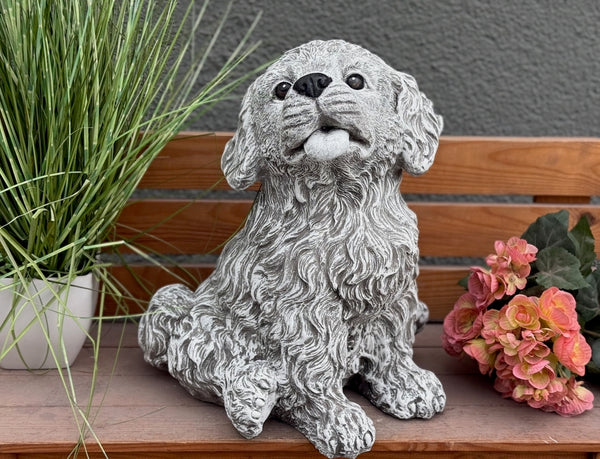 Steinfigur Hund Welpe auf Terrasse – charmanter sitzender Lockenfell-Welpe als frostfeste Gartendeko, handgefertigt in Duisburg mit Blumen im Hintergrund