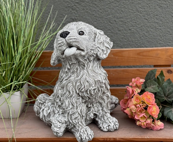 Steinfigur Hund Welpe auf Terrasse – charmanter sitzender Lockenfell-Welpe als frostfeste Gartendeko, handgefertigt in Duisburg mit Blumen im Hintergrund